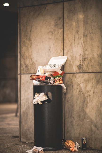 A black cylindrical litter bin with a rounded lid is positioned against a large, beige-toned wall made of smooth, rectangular stone tiles. The lid is open, revealing a collection of discarded food pac