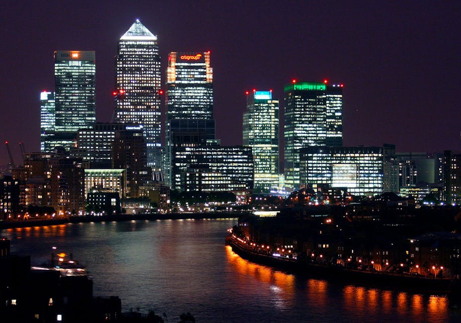 A nighttime cityscape of Canary Wharf featuring several illuminated skyscrapers with modern glass facades. Prominent buildings include the One Canada Square tower with its pyramid-shaped roof, and oth