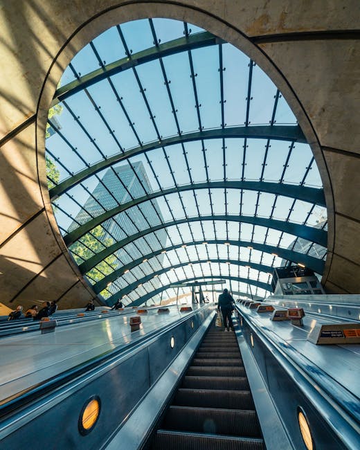 A streamlined red London Underground train is moving along an elevated track above a river in an urban setting. The train features a modern design with smooth exterior panels and large window sections